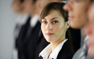 Young woman standing in line with co-workers. She is the only one in focus. She is looking to her left at the camera with a slight smile.
