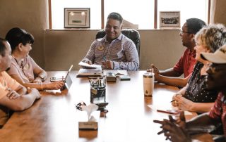 A group of men and women of various ages and ethnicities sit around a meeting table.