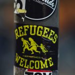 A sticker on a pole that says "Refugees welcome" and has a drawing of the silhouettes of two adults and a child fleeing.